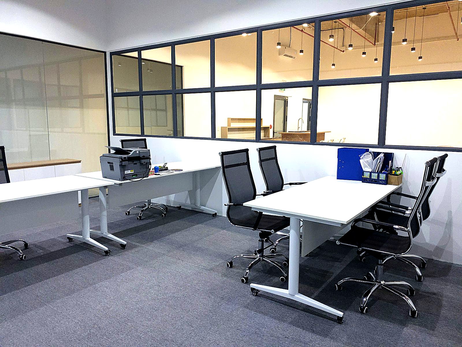 Dedicated desks with mesh chairs beside glass-walled private offices on grey carpet.